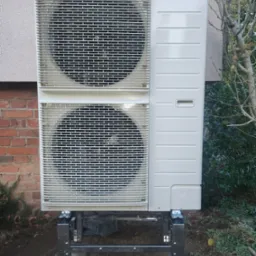 Zewnętrzna jednostka klimatyzacyjna Panasonic zamontowana na metalowej konstrukcji wsporczej, umieszczona przy ścianie budynku z cegły i elementami elewacji, otoczona zielenią.