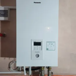 Biały, wiszący na ścianie moduł pompy ciepła Panasonic Aquarea Smart Cloud z widocznymi przyłączami hydraulicznymi i wyświetlaczem pokazującym temperaturę, zamontowany w pomieszczeniu z kafelkami...