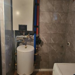 ELNIKO Angelika Strzyż - Biały bojler elektryczny Galmet i wiszący piec Innova z podłączeniami hydraulicznymi na tle marmurowych płytek w łazience.