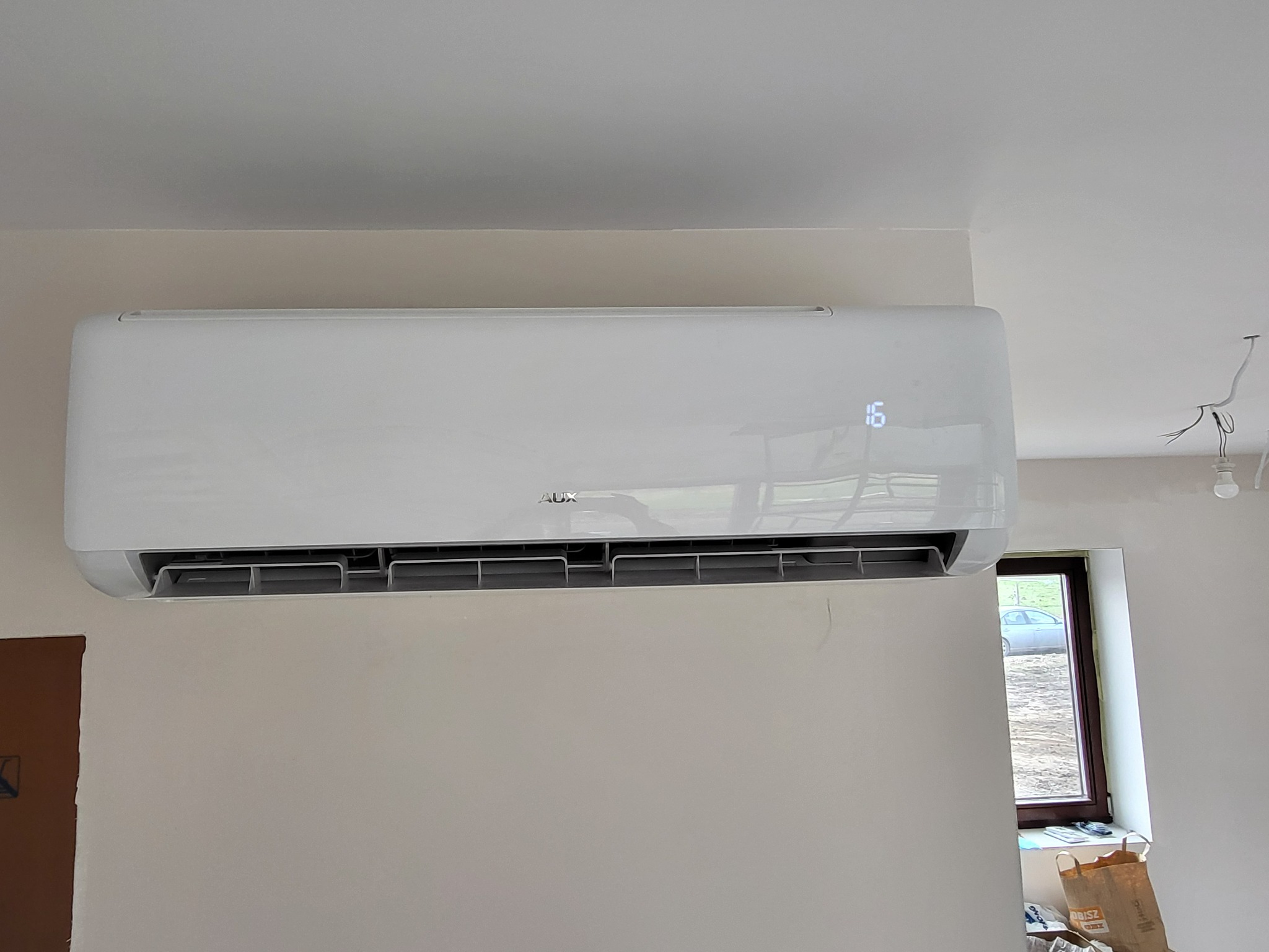 Zamontowany biały klimatyzator AUX z wyświetlaczem temperatury 16 stopni w domu w Prochowicach, widoczne niedokończone wykończenie ściany i okno z widokiem na zewnątrz.
