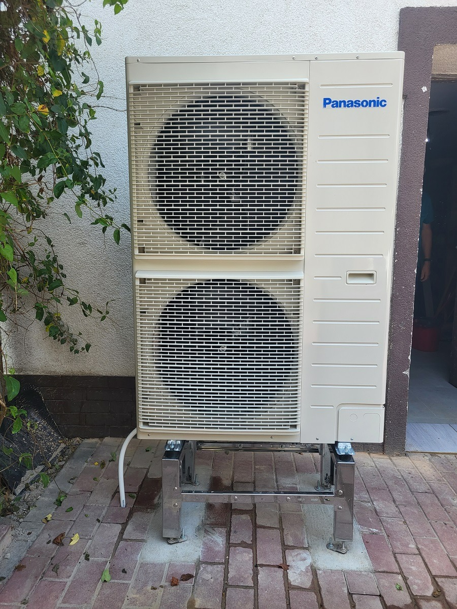 Zewnętrzna jednostka klimatyzacji Panasonic zamontowana na metalowej konstrukcji stojącej na ceglanym chodniku, obok ściany budynku z zielonym pnączem.