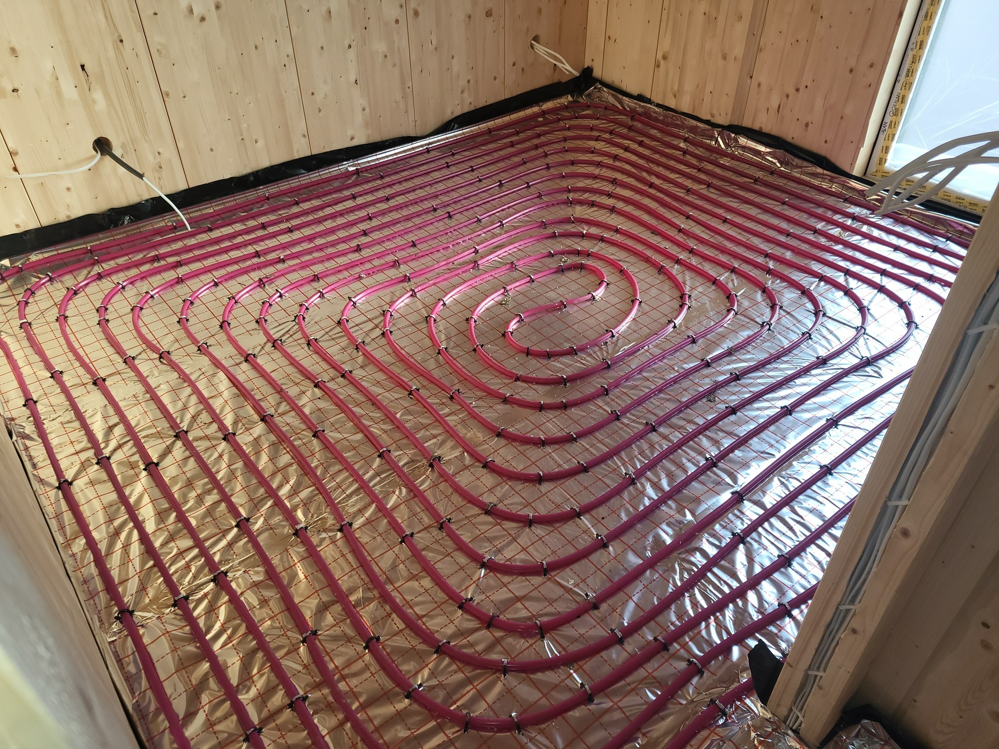 Instalacja ogrzewania podłogowego w spiralnym układzie, rurki w kolorze różowym mocowane do folii aluminiowej na drewnianej podłodze, widoczne kable i fragment okna dachowego.