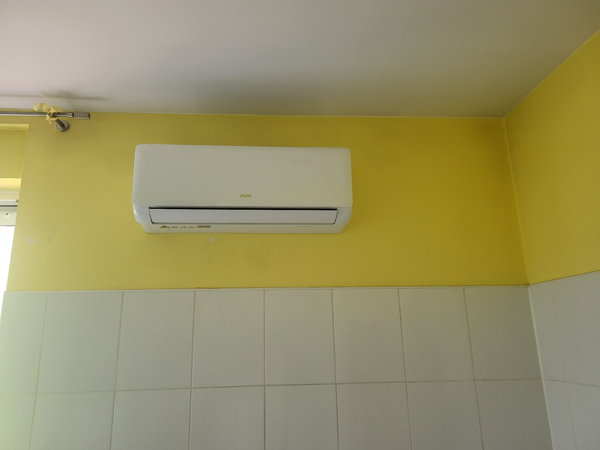 Biały klimatyzator marki AUX zamontowany na żółtej ścianie nad białą, kafelkową ścianą w pomieszczeniu.