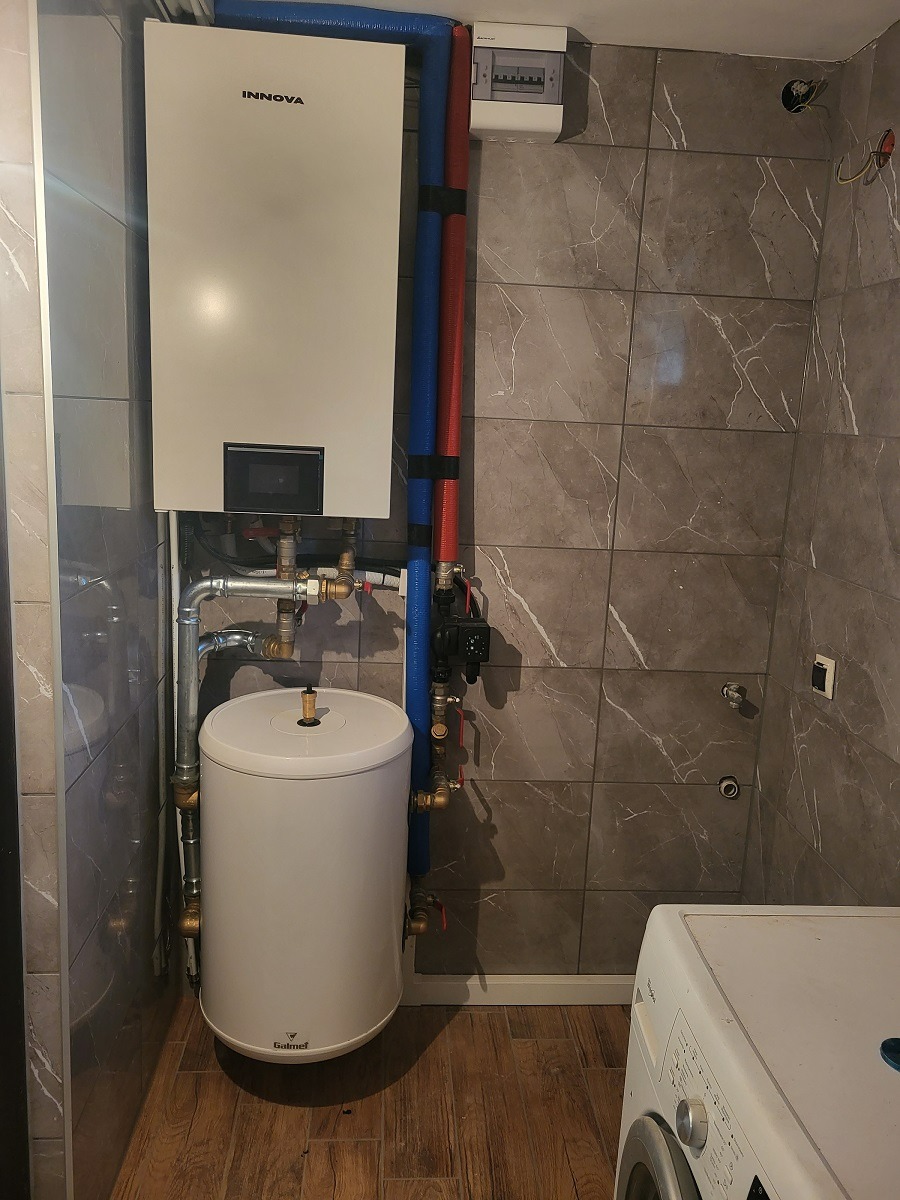 Biały bojler elektryczny Galmet i wiszący piec Innova z podłączeniami hydraulicznymi na tle marmurowych płytek w łazience.