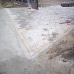 Układanie jasnoszarej kostki brukowej na posesji, widoczne narzędzia pracy i niedokończone obrzeże z jasnych kostek.