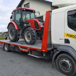 JGauto - Pomoc Drogowa Holowanie 24h Jędrzejów - Czerwony traktor Zetor zabezpieczony pasami na platformie lawety z napisem 'Pomoc Drogowa' na tle domu i pola.