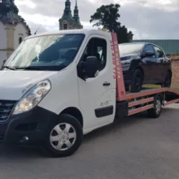 Biała laweta Renault z czarnym samochodem osobowym na platformie, zaparkowana na ulicy z zabytkowym kościołem w tle.