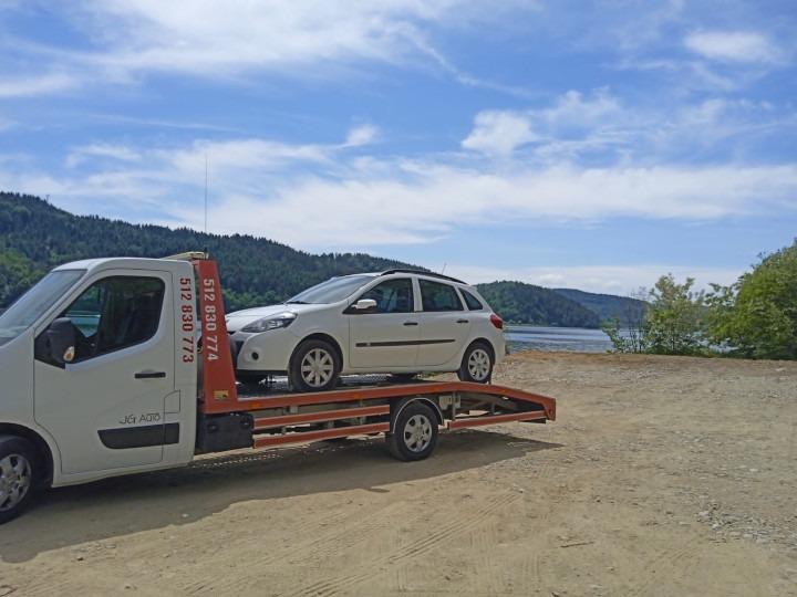 Biała laweta z białym samochodem osobowym na tle jeziora i zalesionych wzgórz w słoneczny dzień.