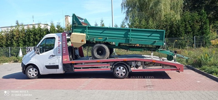 Biała laweta z zieloną przyczepą rolniczą na platformie, widok z boku na tle zieleni i kostki brukowej.