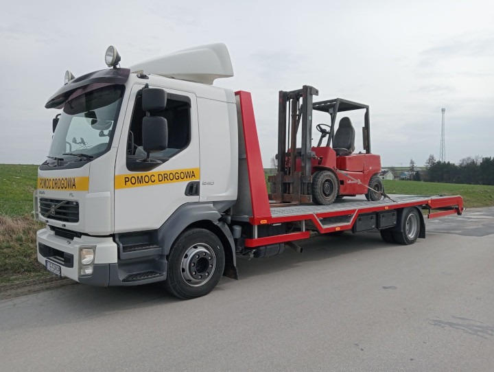 Biała laweta Volvo z napisem 'Pomoc Drogowa' przewozi czerwony wózek widłowy na platformie, zabezpieczony linami. W tle widać zielone pole i słup telekomunikacyjny.