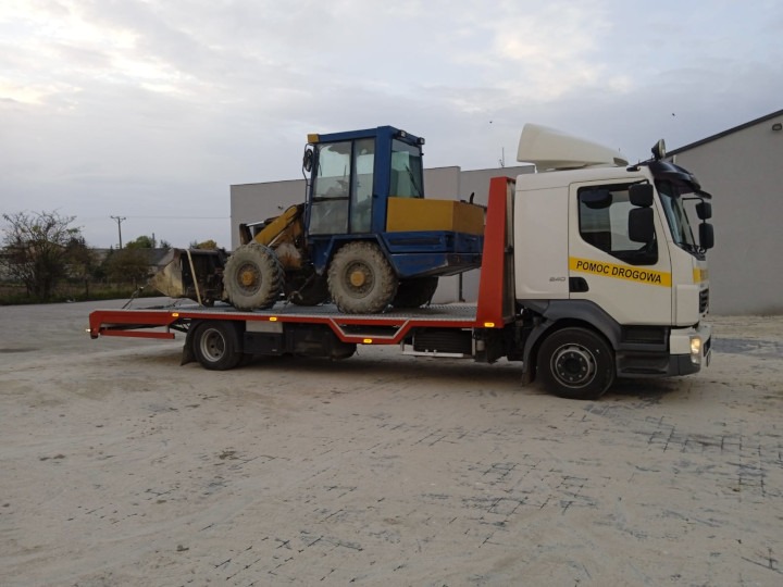 Laweta z napisem 'Pomoc Drogowa' przewozi małą ładowarkę kołową w kolorach niebieskim i żółtym na placu manewrowym.