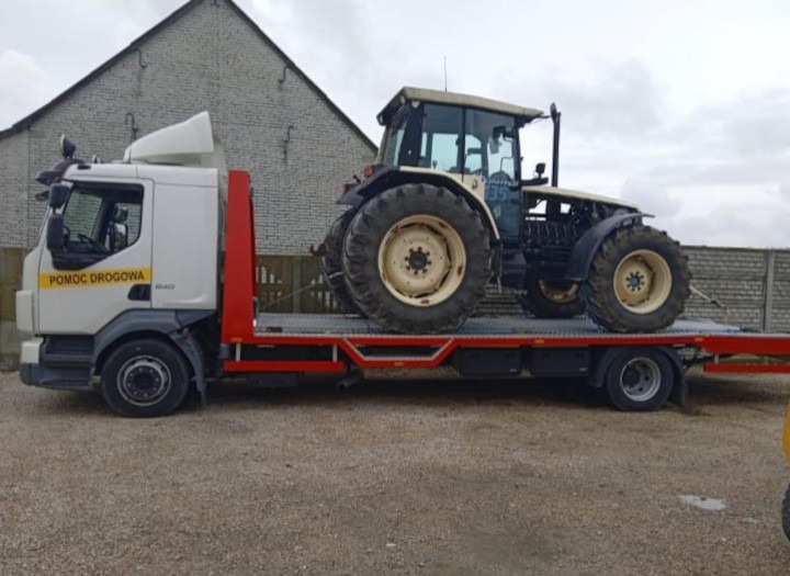 Ciężarówka pomocy drogowej z platformą transportową przewożąca traktor rolniczy z kabiną na tle murowanego budynku.