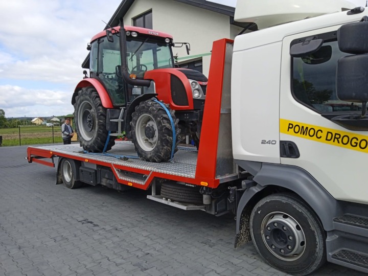 Czerwony traktor Zetor zabezpieczony pasami na platformie lawety z napisem 'Pomoc Drogowa' na tle domu i pola.
