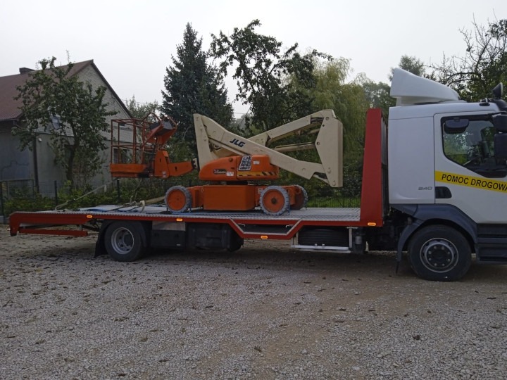 Pomoc drogowa z platformą załadunkową przewozi dwie pomarańczowe platformy robocze JLG na tle drzew i budynku.