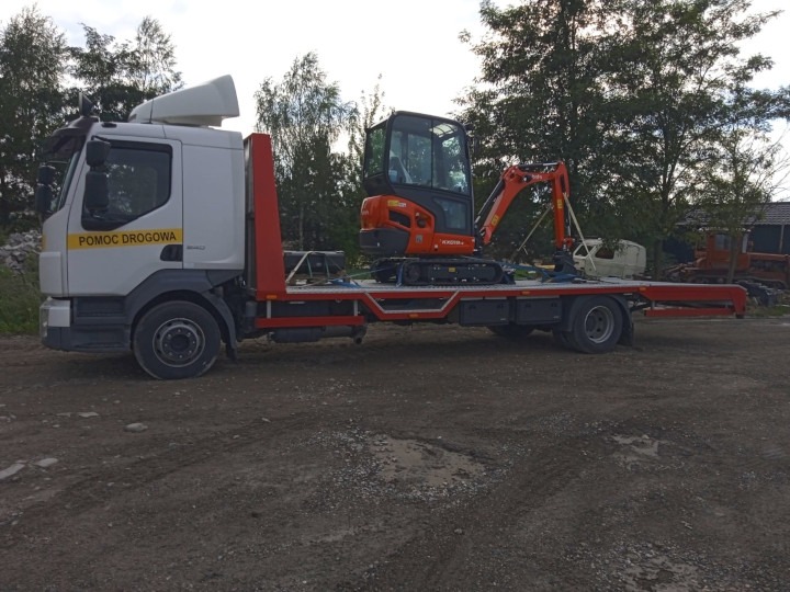 Biała ciężarówka z napisem 'POMOC DROGOWA' przewozi pomarańczową mini koparkę na czerwonej platformie, na tle drzew i ziemi.