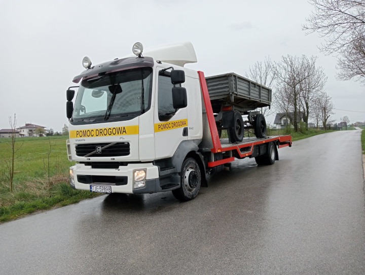 Biała laweta Volvo z żółtym napisem 'Pomoc Drogowa' przewozi przyczepę na mokrej, asfaltowej drodze w pochmurny dzień. Widoczne zabudowania w tle.
