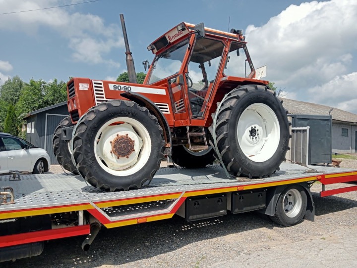 Pomarańczowy traktor Fiat 90-90 umieszczony na platformie lawety, zabezpieczony pasami, z widocznymi detalami opon i kabiny, na tle błękitnego nieba z chmurami.