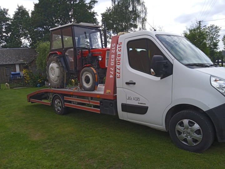 Czerwony traktor Zetor na platformie białego samochodu pomocy drogowej z widocznym numerem telefonu, na tle zielonego trawnika i budynków gospodarczych.