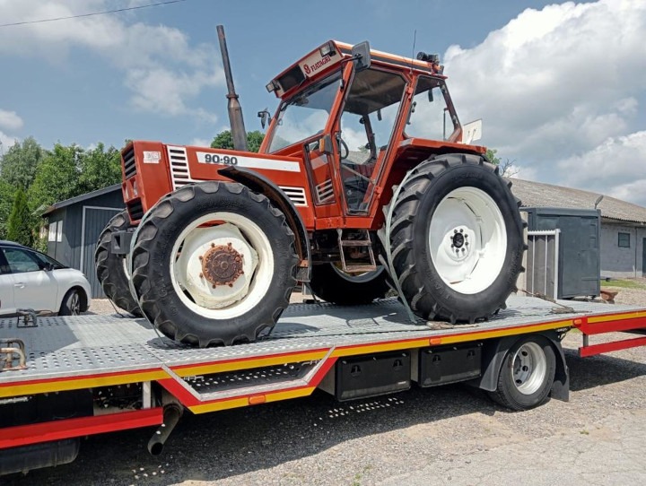 Czerwony ciągnik rolniczy Fiat 90-90 zabezpieczony pasami na platformie niskopodwoziowej z żółto-czerwonymi elementami, widoczny z boku, z budynkiem w tle.