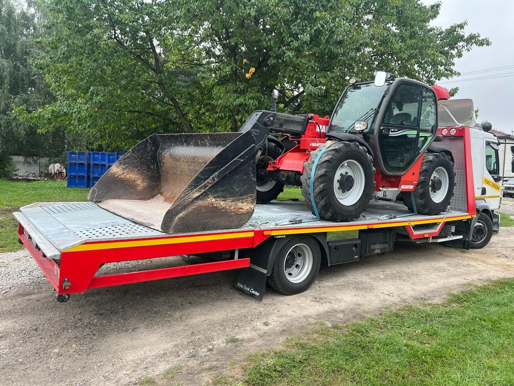Czerwony ładowarka teleskopowa marki Manitou, zabezpieczona niebieskimi pasami, przewożona na platformie lawety z żółto-czerwonymi elementami. W tle widoczne drzewa i niebieskie kontenery.