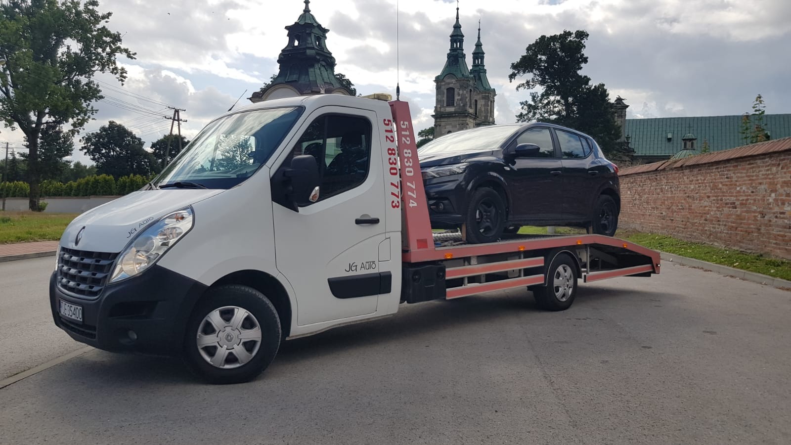 Czarny samochód osobowy na lawecie w Jędrzejowie, z widocznym numerem telefonu na nadwoziu, na tle zabytkowego kościoła.
