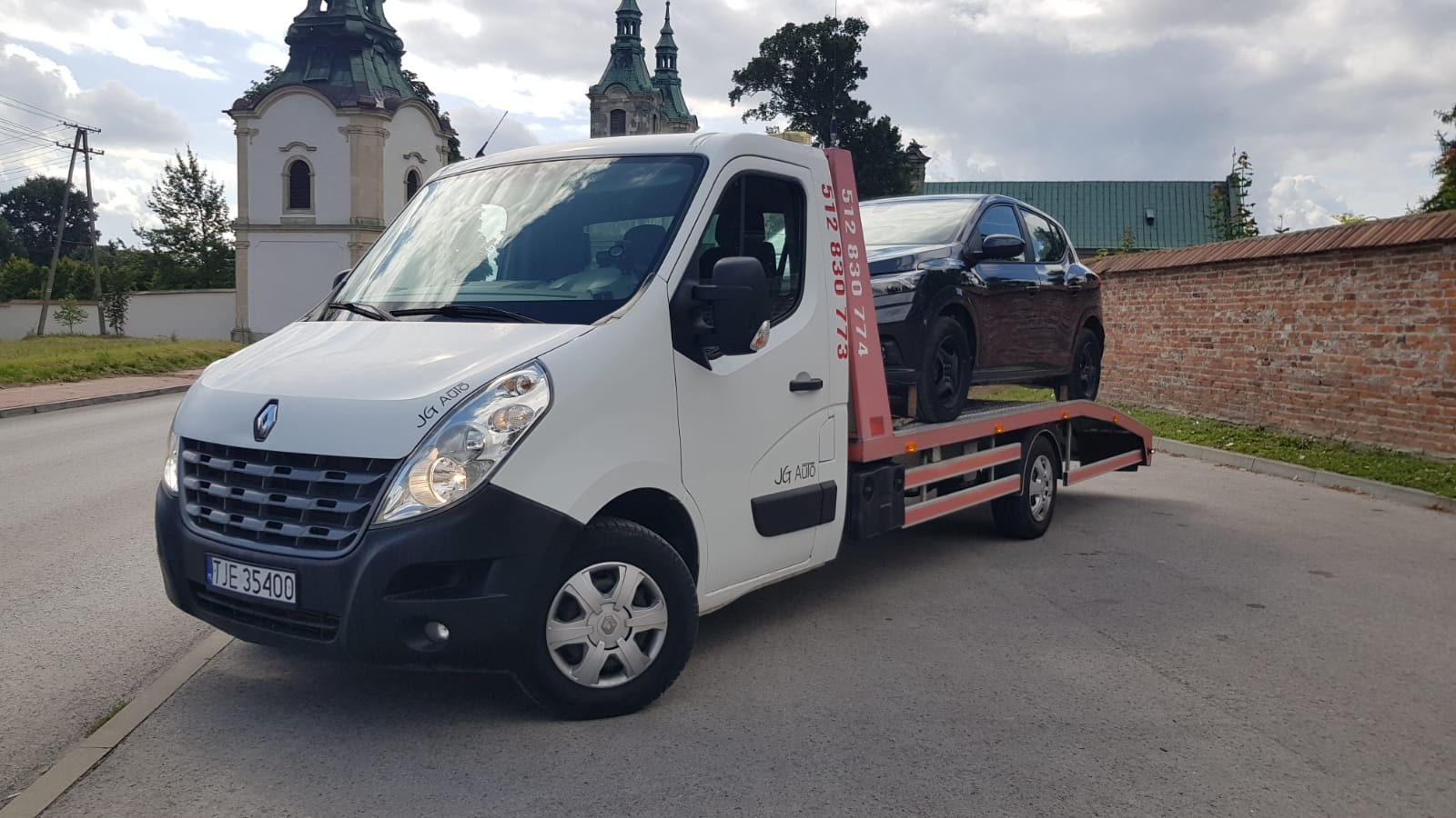 Biała laweta Renault z czarnym samochodem osobowym na platformie, zaparkowana na ulicy z zabytkowym kościołem w tle.