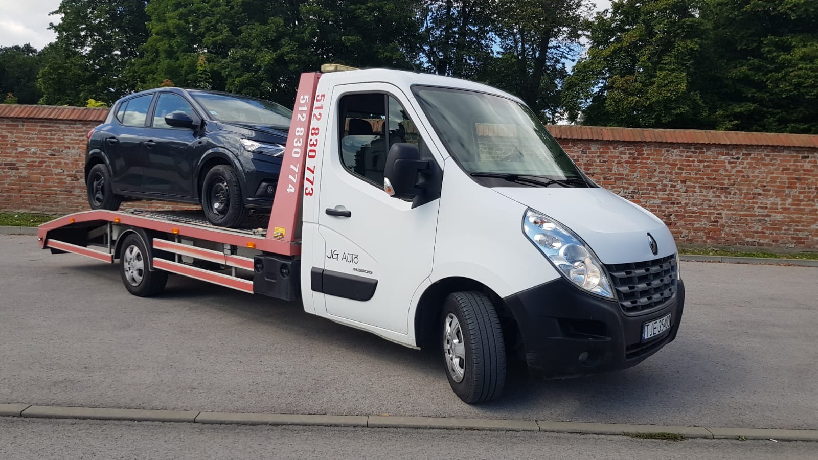 Biała laweta Renault z czarnym samochodem osobowym na platformie, zaparkowana na asfaltowej drodze przed ceglanym murem i zielonymi drzewami.