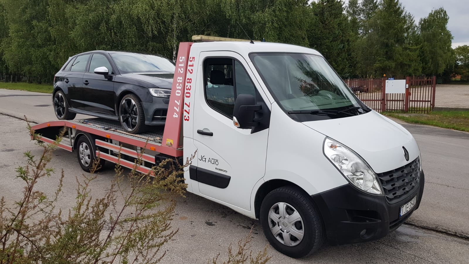 Biała laweta z czarnym Audi na platformie, zaparkowana na poboczu drogi z widocznym numerem telefonu na czerwonym słupku platformy.