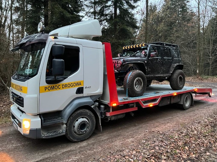 Czarny, terenowy Jeep Wrangler na lawecie pomocy drogowej w lesie, widoczne błoto na samochodzie i otoczeniu.