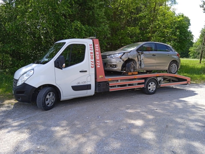 Biała laweta z uszkodzonym srebrnym samochodem osobowym na platformie, widoczne drzewa w tle.