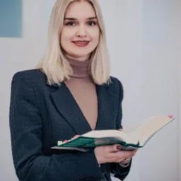 Uśmiechnięta blondynka w szarym żakiecie i golfie, trzyma otwarta księgę z kolorowymi zakładkami na tle jasnej ściany.