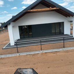 Mistral - A Zarzycki Sp. Jawna - Nowoczesny, parterowy dom z płaskim dachem, czarną stolarką okienną i wykończonym tarasem z kostki brukowej, ogrodzony siatką na betonowej podmurówce, w trakcie zagospodarowania terenu.