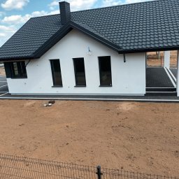 Mistral - A Zarzycki Sp. Jawna - Nowoczesny, parterowy dom z ciemną dachówką i czarnymi oknami, widoczny z góry, z niezagospodarowanym terenem wokół i fragmentem ogrodzenia z siatki na pierwszym planie.