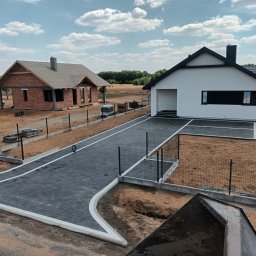 Mistral - A Zarzycki Sp. Jawna - Widok z góry na nowo wybudowany dom jednorodzinny z białymi ścianami i czarnym dachem, szara kostka brukowa na podjeździe z białymi obrzeżami, w tle dom w trakcie budowy z czerwonej cegły...