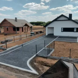 Widok z góry na nowo wybudowany dom jednorodzinny z białymi ścianami i czarnym dachem, szara kostka brukowa na podjeździe z białymi obrzeżami, w tle dom w trakcie budowy z czerwonej cegły...