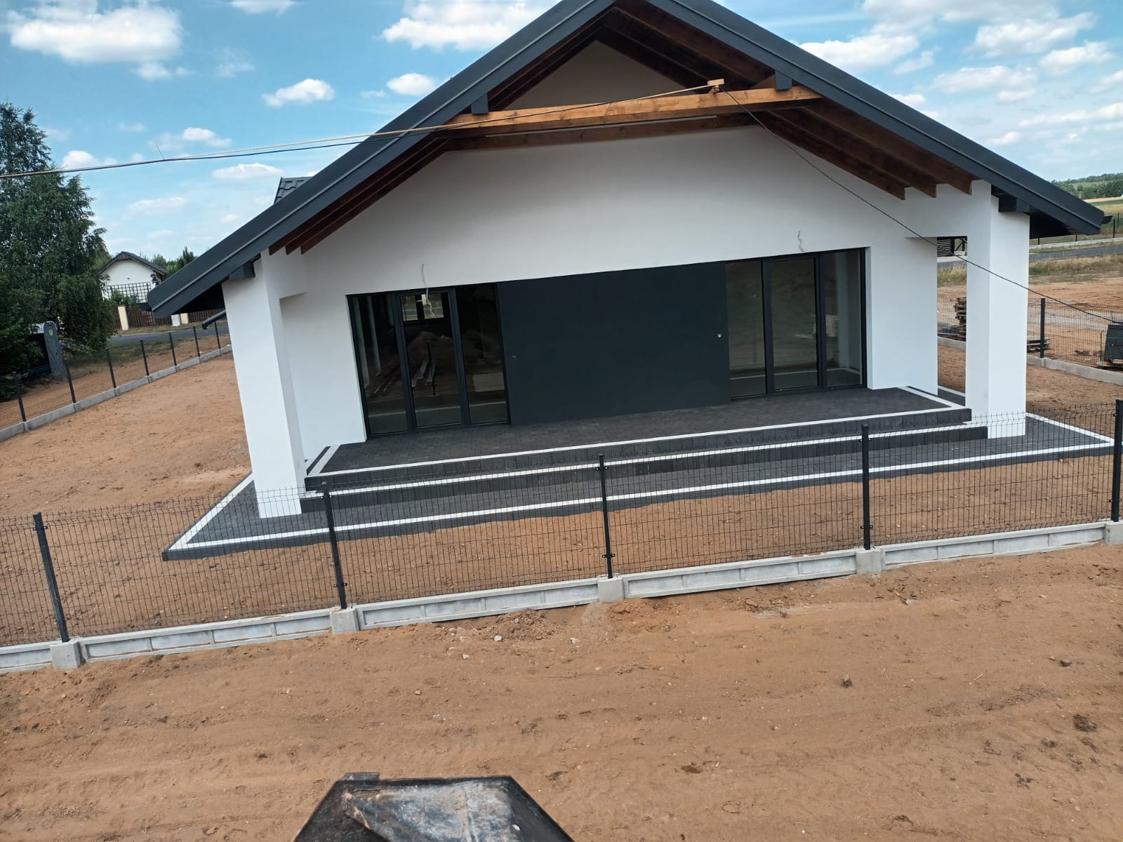 Nowoczesny, parterowy dom z płaskim dachem, czarną stolarką okienną i wykończonym tarasem z kostki brukowej, ogrodzony siatką na betonowej podmurówce, w trakcie zagospodarowania terenu.