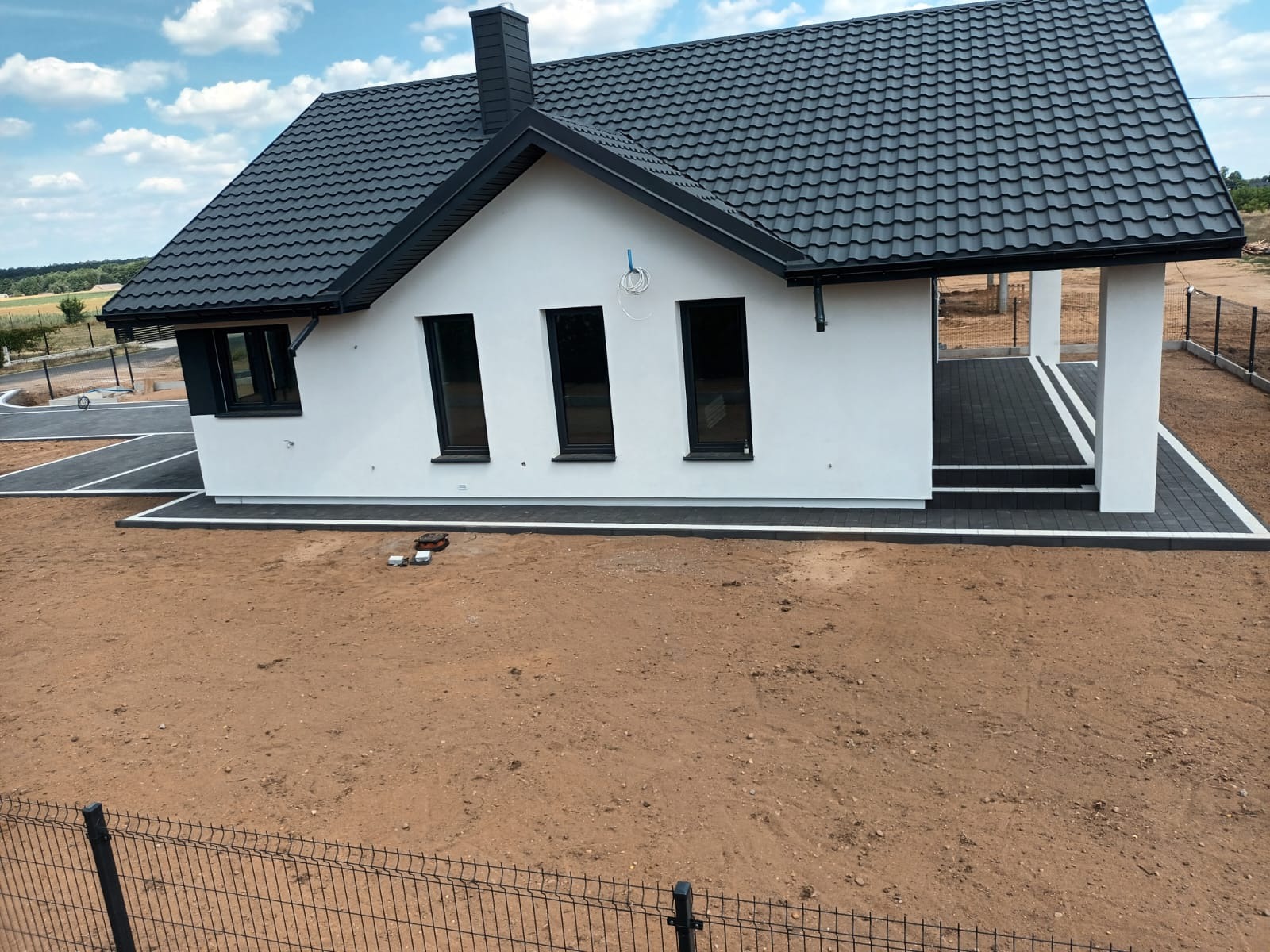 Nowoczesny, parterowy dom z ciemną dachówką i czarnymi oknami, widoczny z góry, z niezagospodarowanym terenem wokół i fragmentem ogrodzenia z siatki na pierwszym planie.