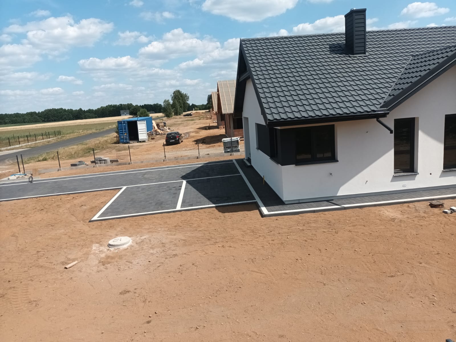 Nowoczesny dom jednorodzinny z szarą kostką brukową na podjeździe i wokół budynku, w tle widoczny kontener i inne domy w trakcie budowy.