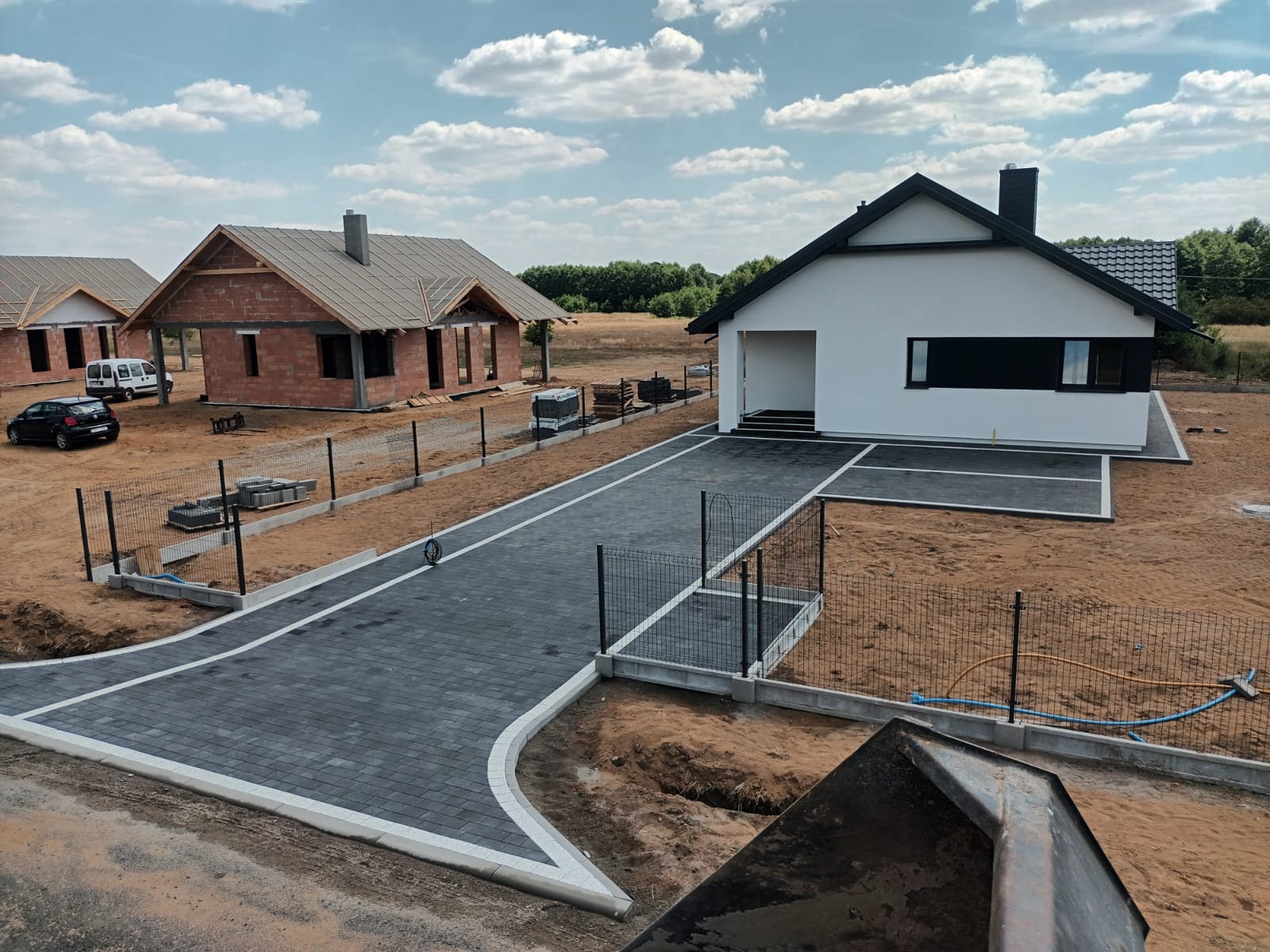 Widok z góry na nowo wybudowany dom jednorodzinny z białymi ścianami i czarnym dachem, szara kostka brukowa na podjeździe z białymi obrzeżami, w tle dom w trakcie budowy z czerwonej cegły...