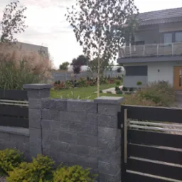 Nowoczesny ogród z domu z szarą, kamienną bramą i ogrodzeniem z metalowymi elementami, prowadzącą do zadbanego trawnika z drzewami i rabatami kwiatowymi.