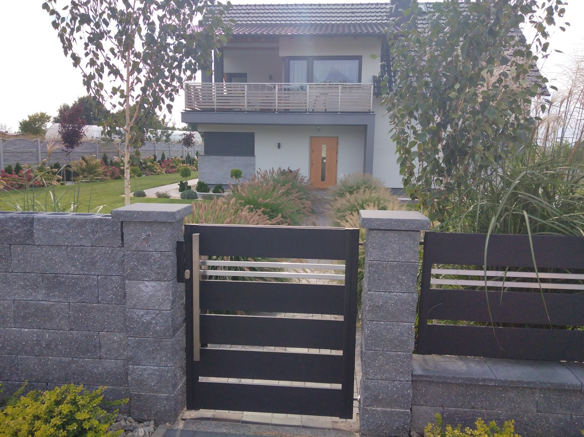 Nowoczesna brama wjazdowa z poziomymi przęsłami i kamiennymi słupkami na tle zadbanego ogrodu z trawami ozdobnymi i dwupiętrowego domu z balkonem.