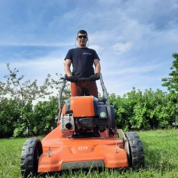 Arbo-Drwal Radosław Zawiślak - Mężczyzna w okularach słonecznych i czarnej koszulce z logo firmy, obsługujący pomarańczową kosiarkę Husqvarna na trawniku w słoneczny dzień.