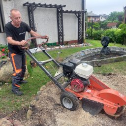 Arbo-Drwal Radosław Zawiślak - Mężczyzna obsługuje frezarkę do pni drzew na posesji z ozdobnym oczkiem wodnym i konstrukcją pergoli
