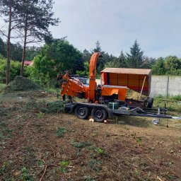 Arbo-Drwal Radosław Zawiślak - Pracownik w pomarańczowym kombinezonie obsługuje pomarańczową rębarkę do gałęzi, obok leżą ścięte gałęzie i pocięte pnie drzew.
