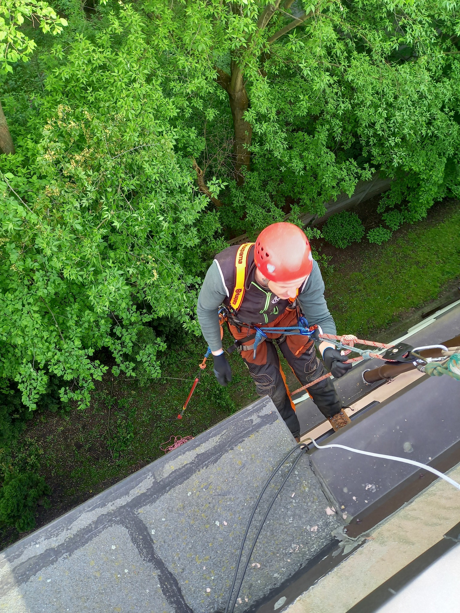 Pracownik w kasku i uprzęży zabezpieczającej, wykonujący prace na wysokości na dachu budynku, widok z góry na tle zieleni.