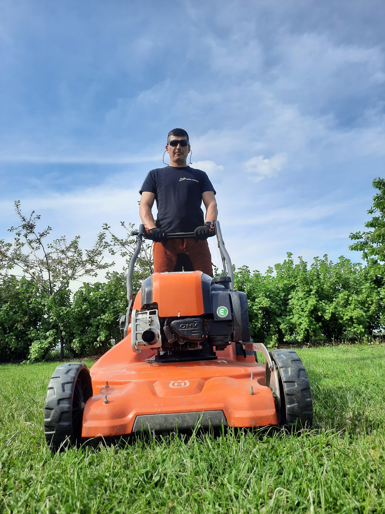 Mężczyzna w okularach słonecznych i czarnej koszulce z logo firmy, obsługujący pomarańczową kosiarkę Husqvarna na trawniku w słoneczny dzień.