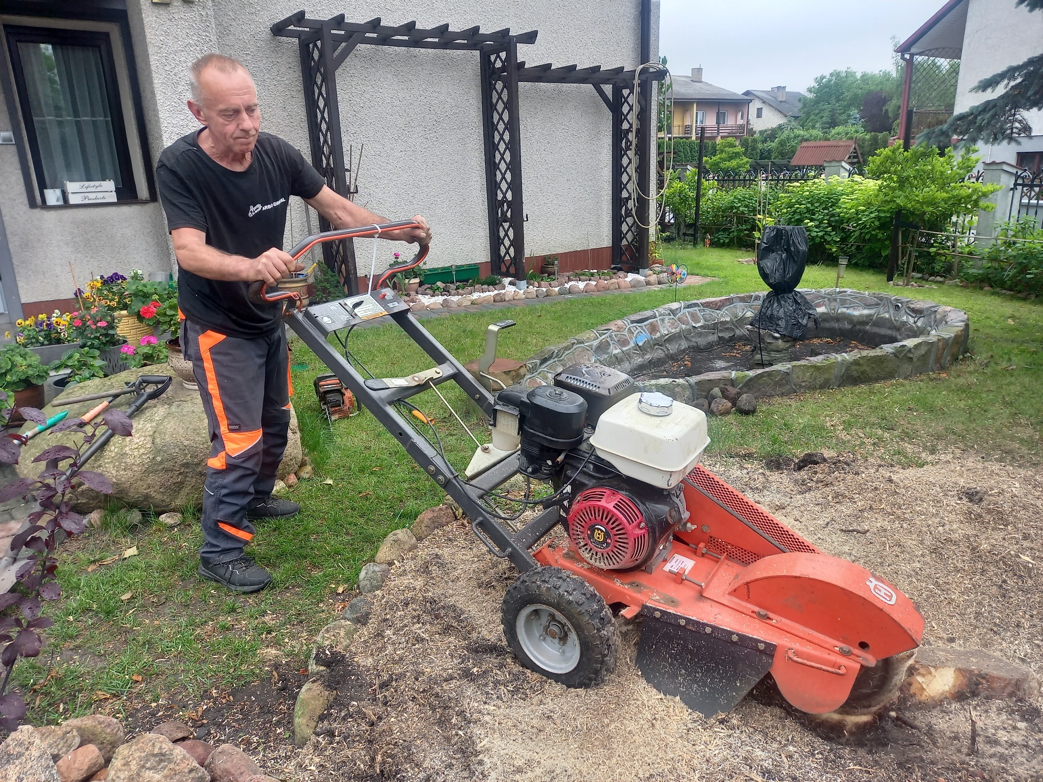 Mężczyzna obsługuje frezarkę do pni drzew na posesji z ozdobnym oczkiem wodnym i konstrukcją pergoli