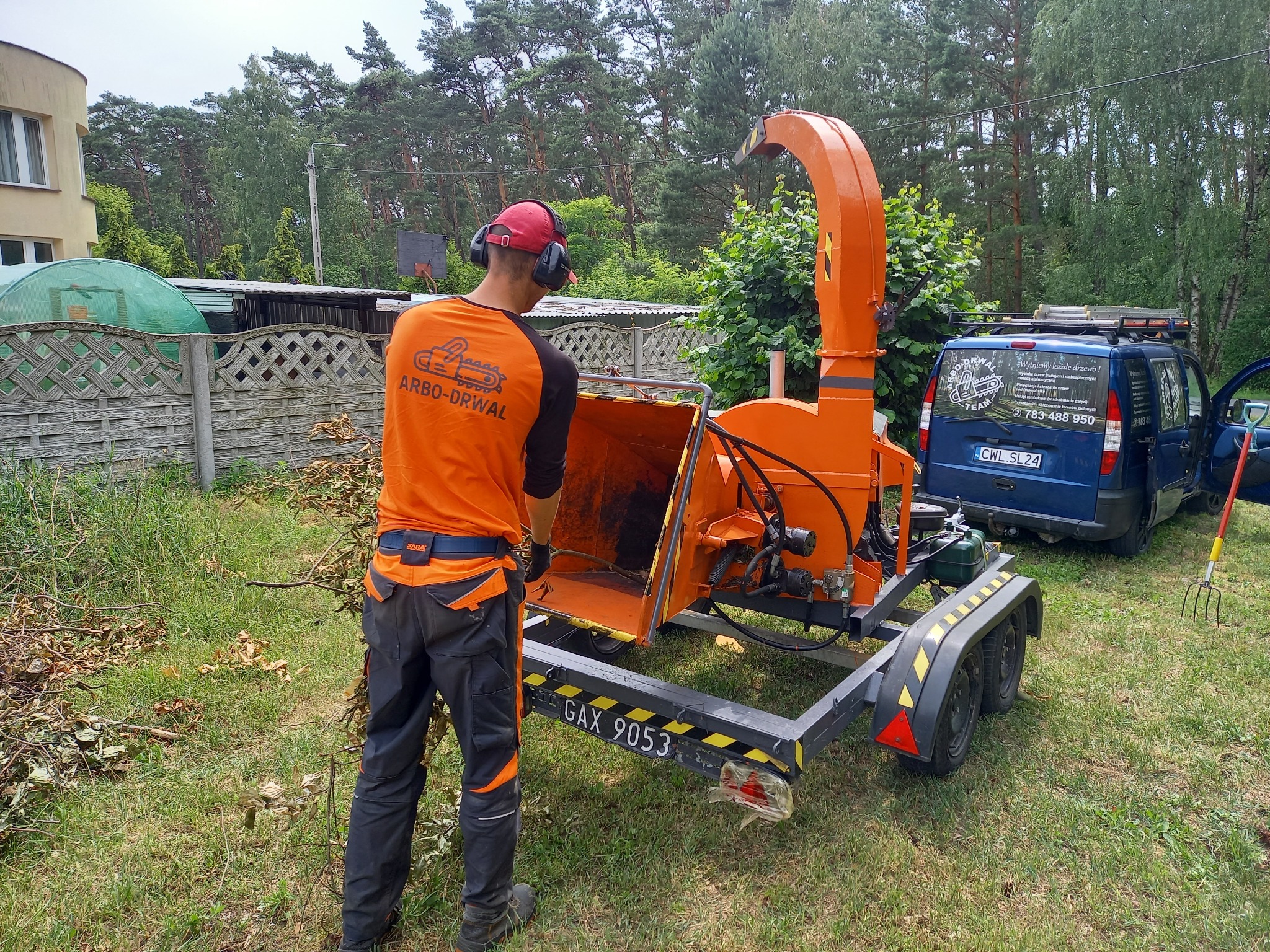 Pracownik w pomarańczowej koszulce z logo firmy Arbo-Drwal, w słuchawkach i czerwonej czapce, obsługuje pomarańczowy rębak do gałęzi na przyczepie, w tle niebieski samochód firmowy z logo i numerem...