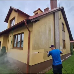 Mężczyzna w niebieskiej koszulce czyści elewację domu z żółtym tynkiem za pomocą myjki ciśnieniowej, widoczny kontrast między czystą a brudną powierzchnią.