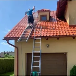 Pracownik na drabinie spryskuje wodą dach z czerwonej dach&oacute;wki na dwukondygnacyjnym domu z beżową elewacją, widoczne ślady czyszczenia.